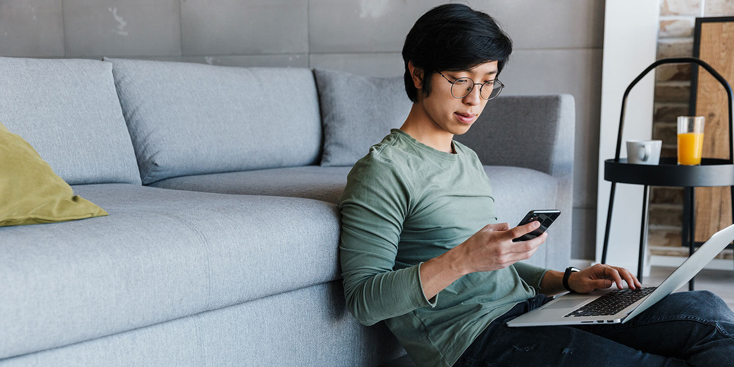 A man sitting on the floor and looking at phone, holding laptop in his lap