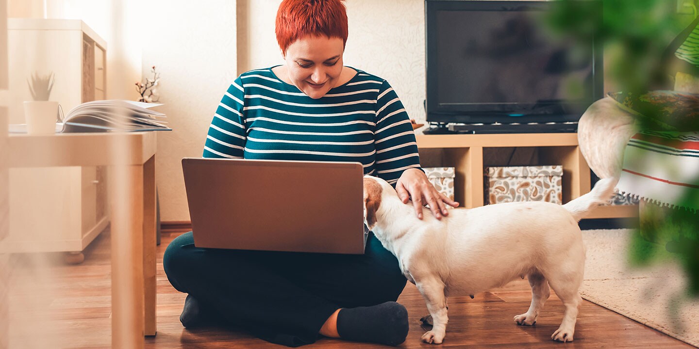 A woman sits on the floor with a notebook computer on her lap, petting a dog.