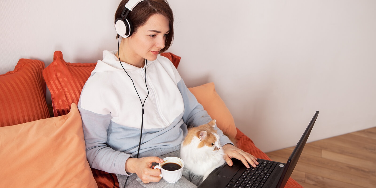 A woman sitting on a couch amd working on laptop wearing headset, with a pet in her lap