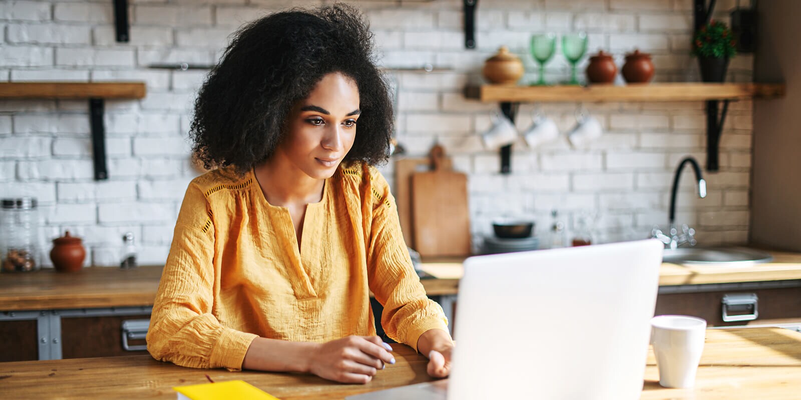 Woman sitting at a table, looking at computer with a coffee cup beside her computer.