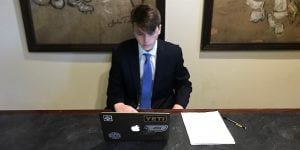 A male college student sitting at a table and looking at a laptop. He is wearing a suit jacket and tie.