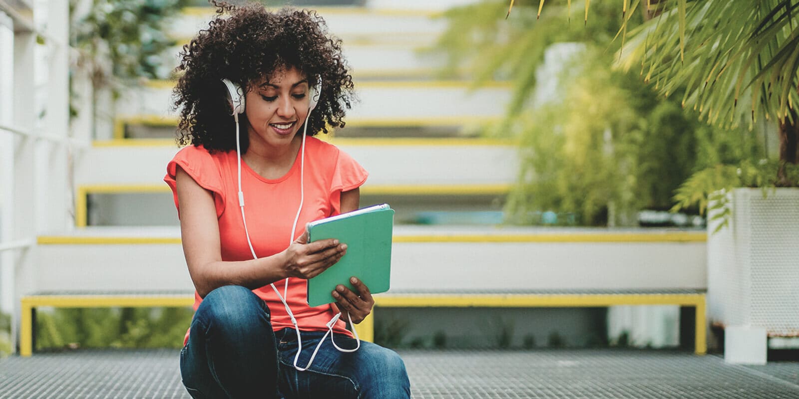 A woman is sitting on some stairs outdoors, wearing headphones and looking at a tablet she is holding.