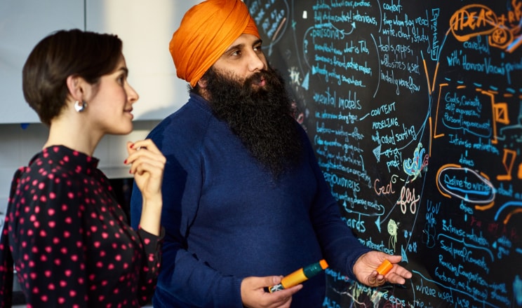 A Sikh man explains ideas on a blackboard to a female colleague.