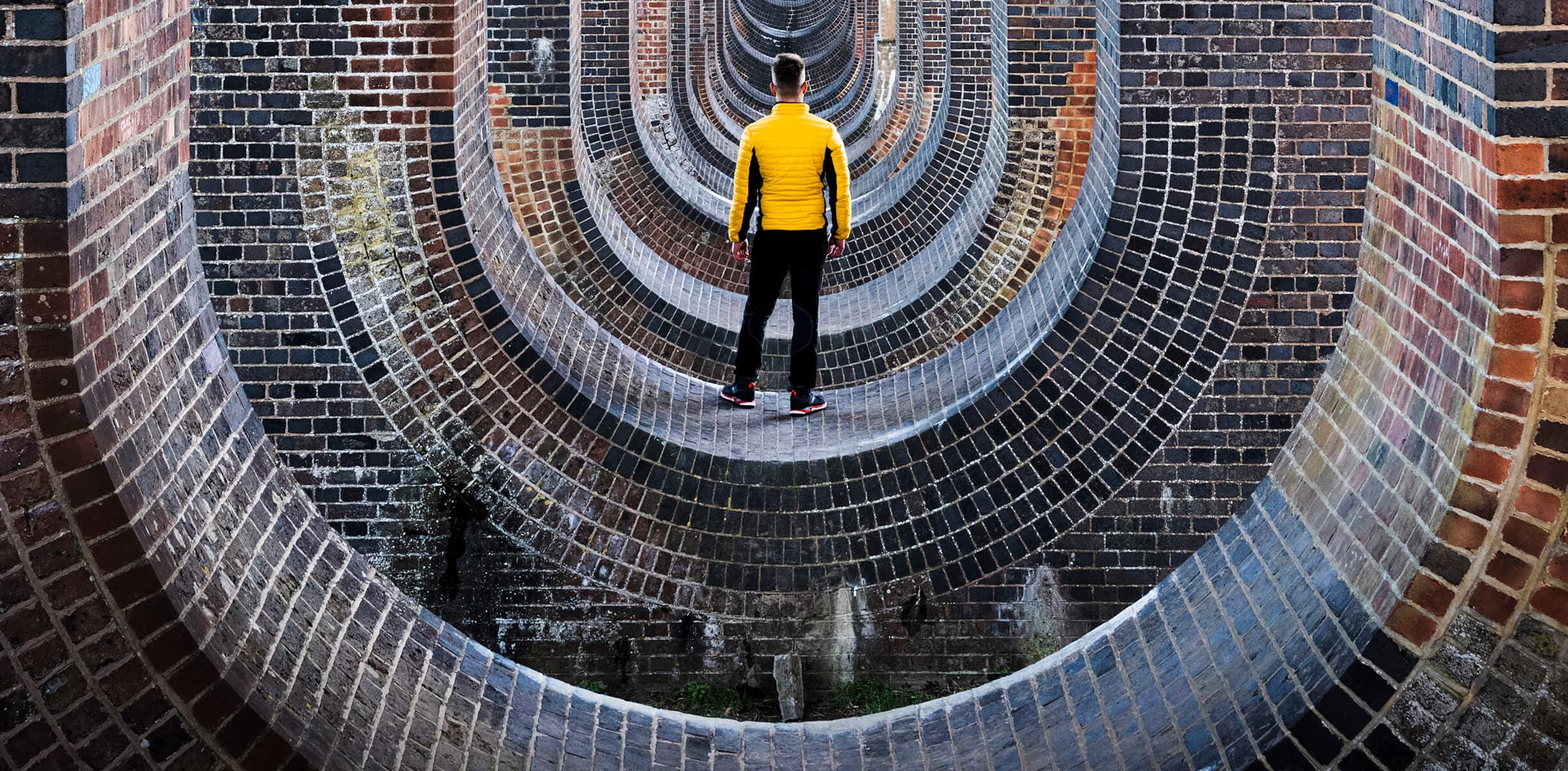 Man in yellow shirt standing within repeating U shaped brick structures. 