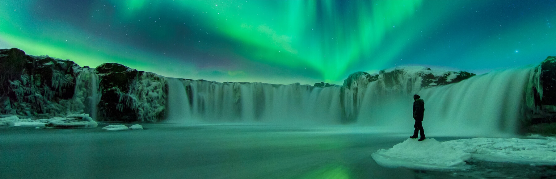 Person on ice looking out at a waterfall under the Northern Lights