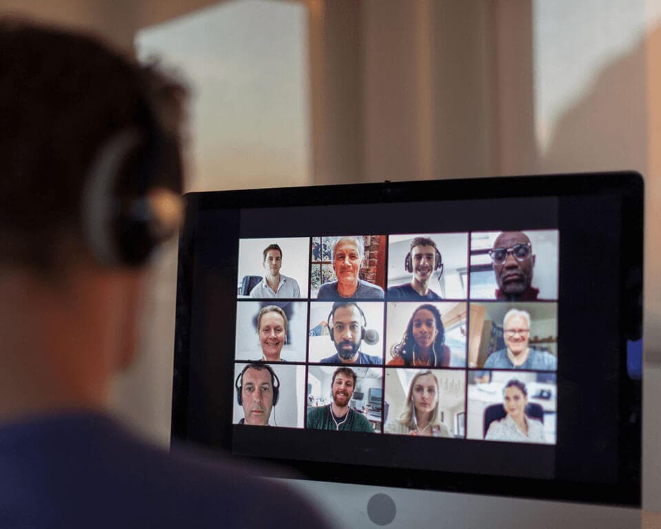 A man wearing headphones is looking at a monitor screen that shows twelve people participating in a video conference call.