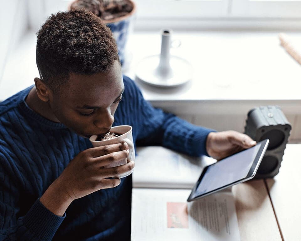 A man drinks out of a mug while looking at a tablet he holds in his hand.