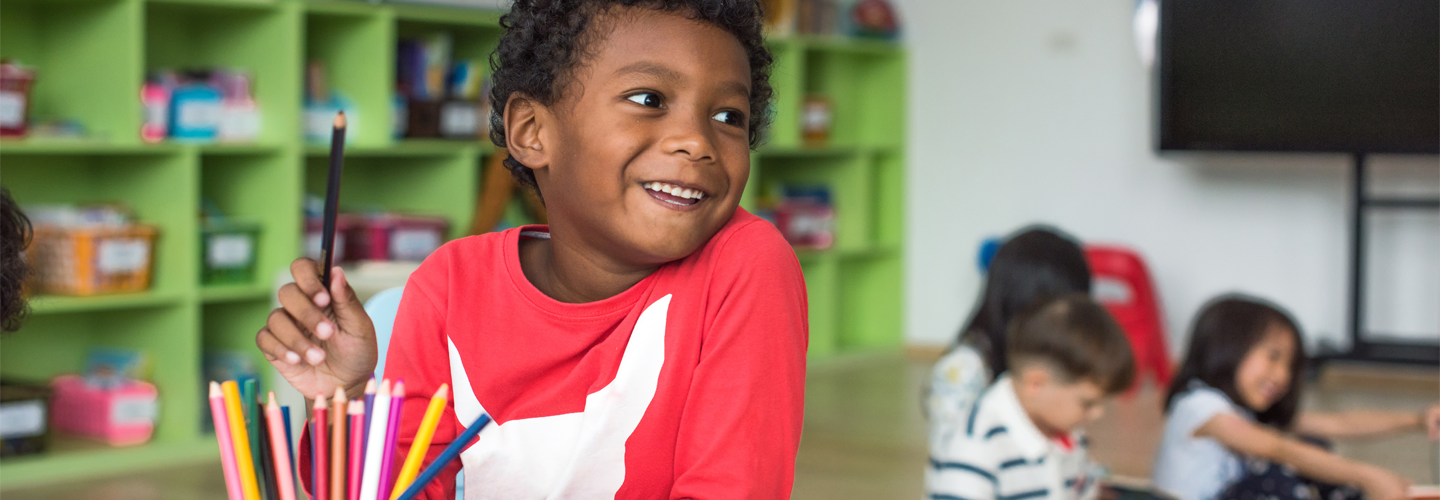 A young child smiling in a classroom with a crayon in his hand.