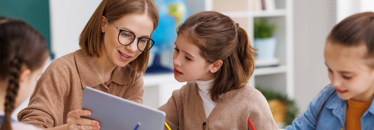 A teacher holding a tablet to a young student in a classroom sat at a table