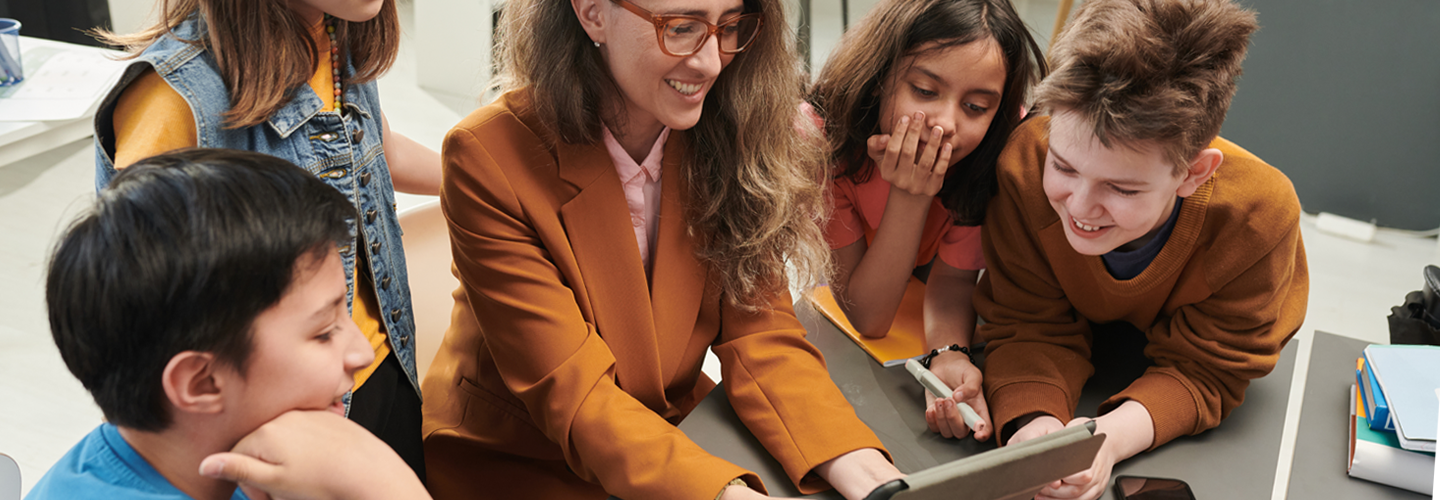 A teacher holding a tablet in a classroom with students around her also looking at the tablet smiling
