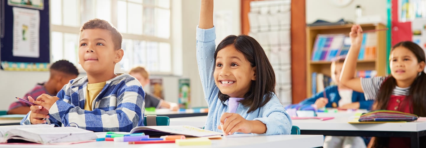 Children sat at desks in a classroom, with one in the middle smiling with her hand up