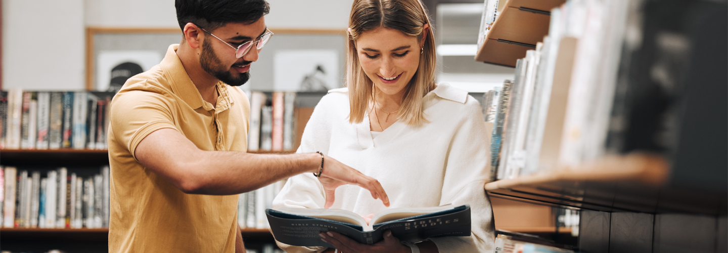Two friends stood over a book in a library reading it and smiling