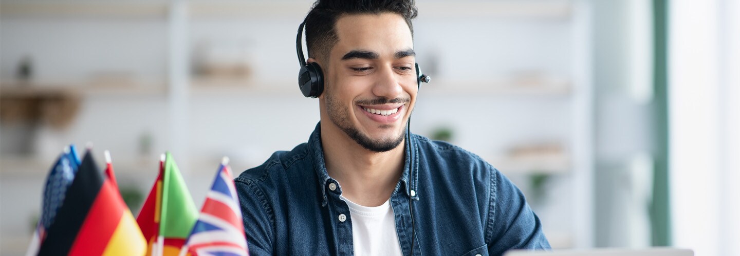 A man with a headset sat at a laptop, with mini flags by him.