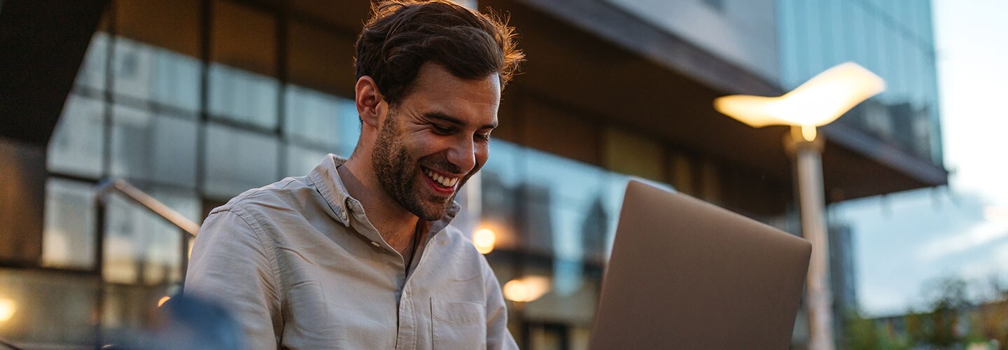 A man sat outside smiling working on a laptop