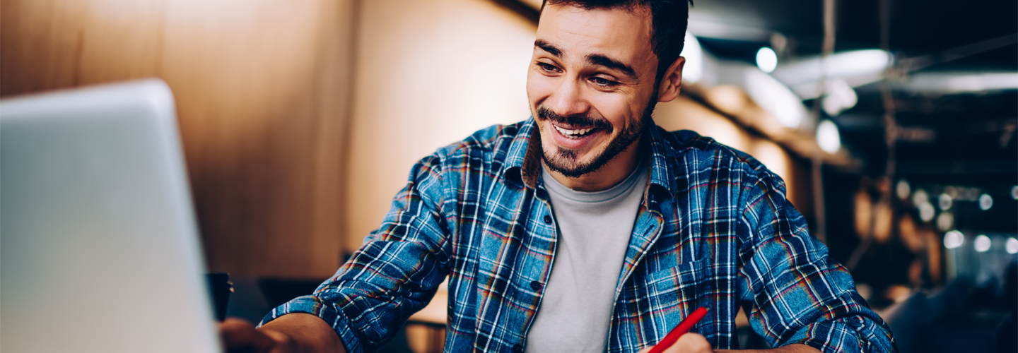 A man looking at a openlaptop with a pen in hand, smiling at the laptop