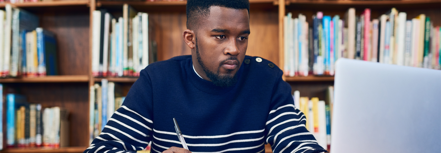 A man sat at a laptop studying in a library holding a pen and paper