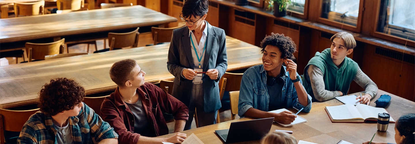 A teacher stood by a long wooden desk where her students are sat smiling at her
