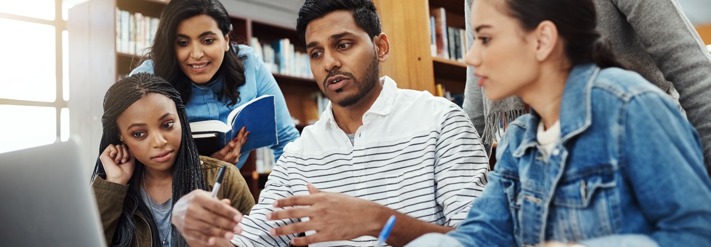 Three individuals are engaged in a discussion around a table in a library, surrounded by books, with one person gesturing with their hands.