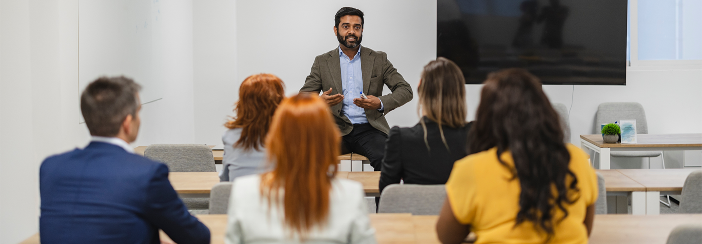A teacher sat in front of his students, who are sat at tables facing him with their backs turned to the camera