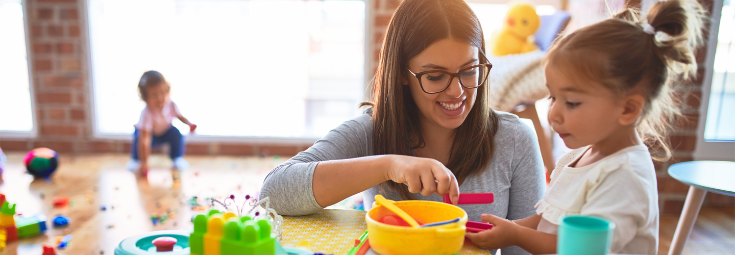 A female teacher sat in a classroom with a young child playing with toys