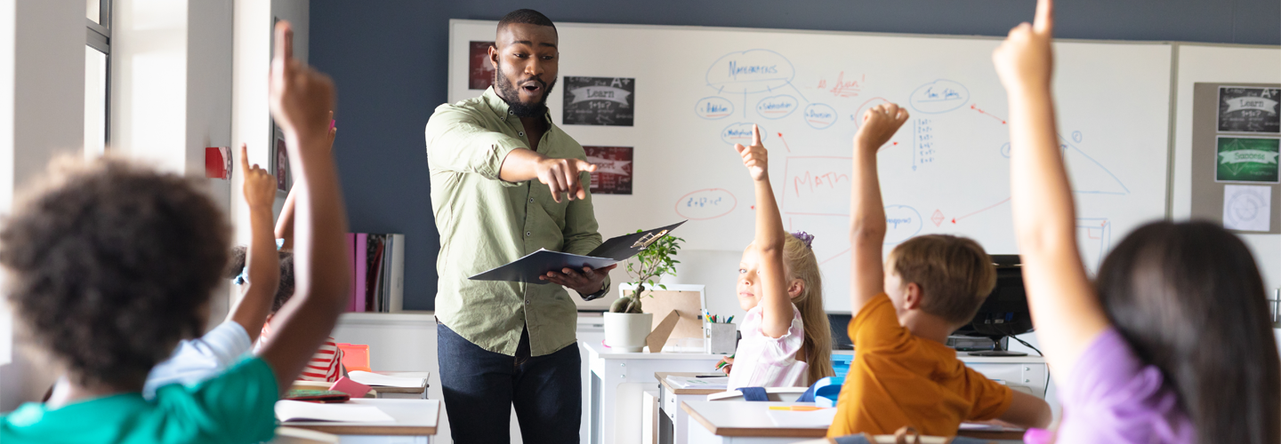 A teacher stood at the front of a class with a book, pointing at a student. Students are sat at desks with their hands raised.
