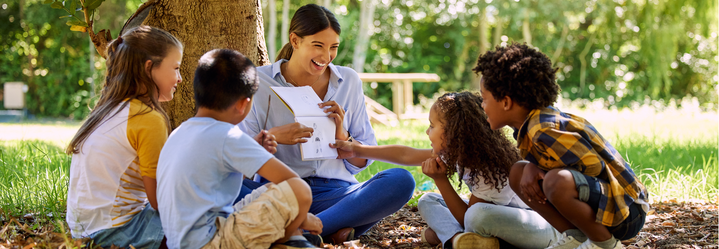 A teacher sat outdoors with young students looking at a book togethr