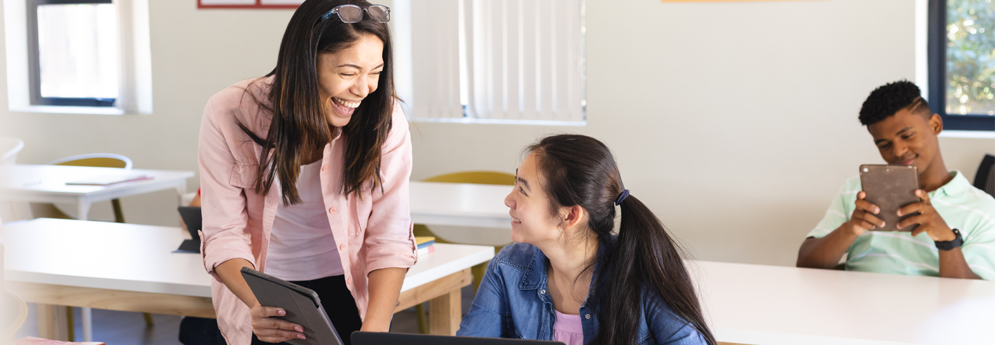 Teacher talking to girl working on laptop in classroom smiling