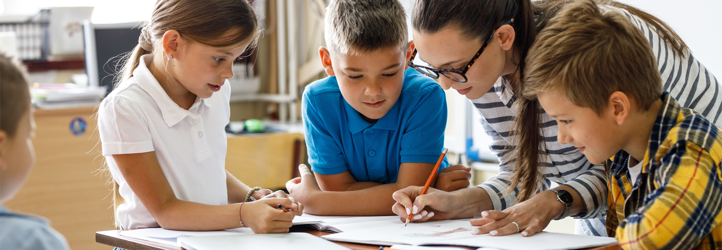 A group of children stood at a table with their teacher watching her write something down on paper