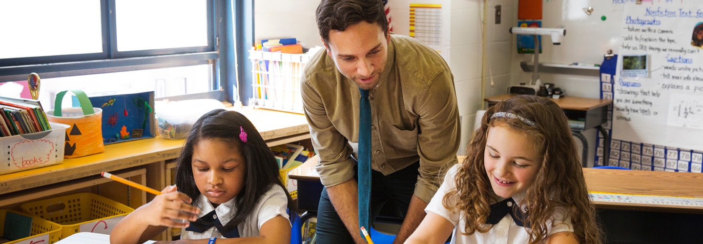a teacher stood with two students sat a desk