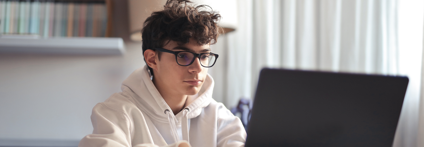 A person in a white hoodie sits at a desk, working on a laptop, with bookshelves in the background.