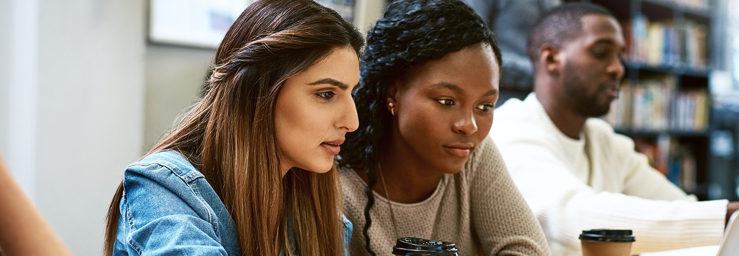 Three students sit at a table in a library, engaged in study with books and a laptop visible.