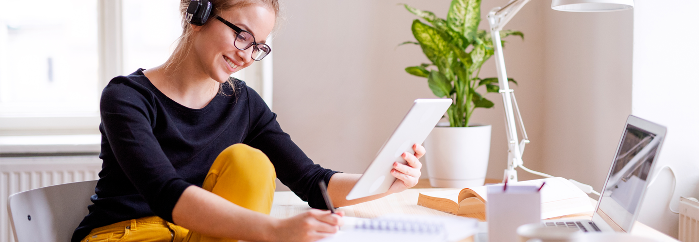 جلست امرأة على مكتب مع جهاز كمبيوتر محمول وجهاز لوحي ودفاتر ملاحظات مفتوحة تدرس