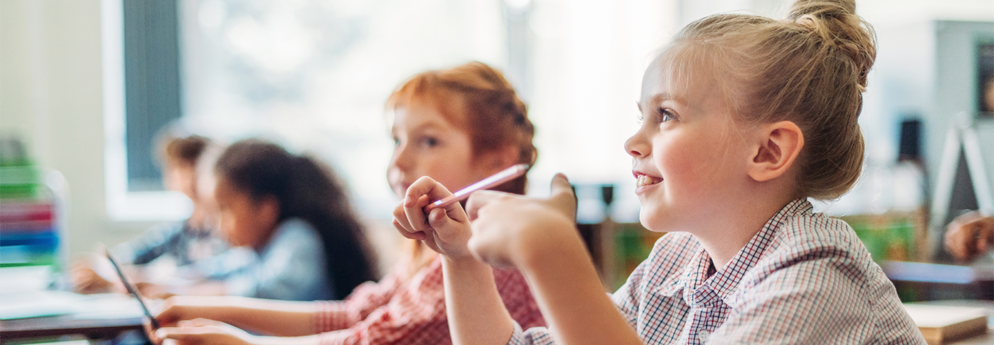 A young girl sat at a desk smiling at the front of the class, with other students in the background.