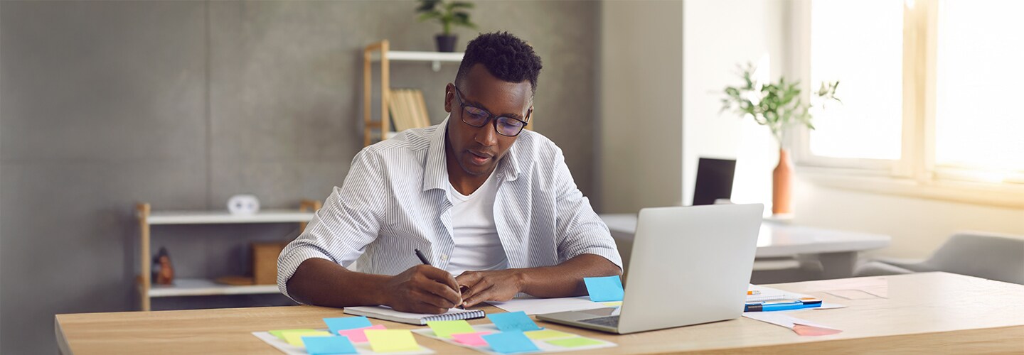 رجل محترف يجلس على مكتب ، ويكتب الملاحظات مع جهاز كمبيوتر محمول مفتوح أمامه.