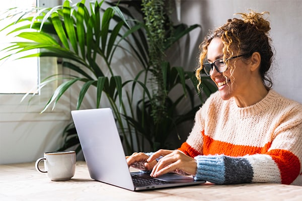 A person in a colorful sweater typing on a laptop at a table near a window with a potted plant and a coffee mug.
