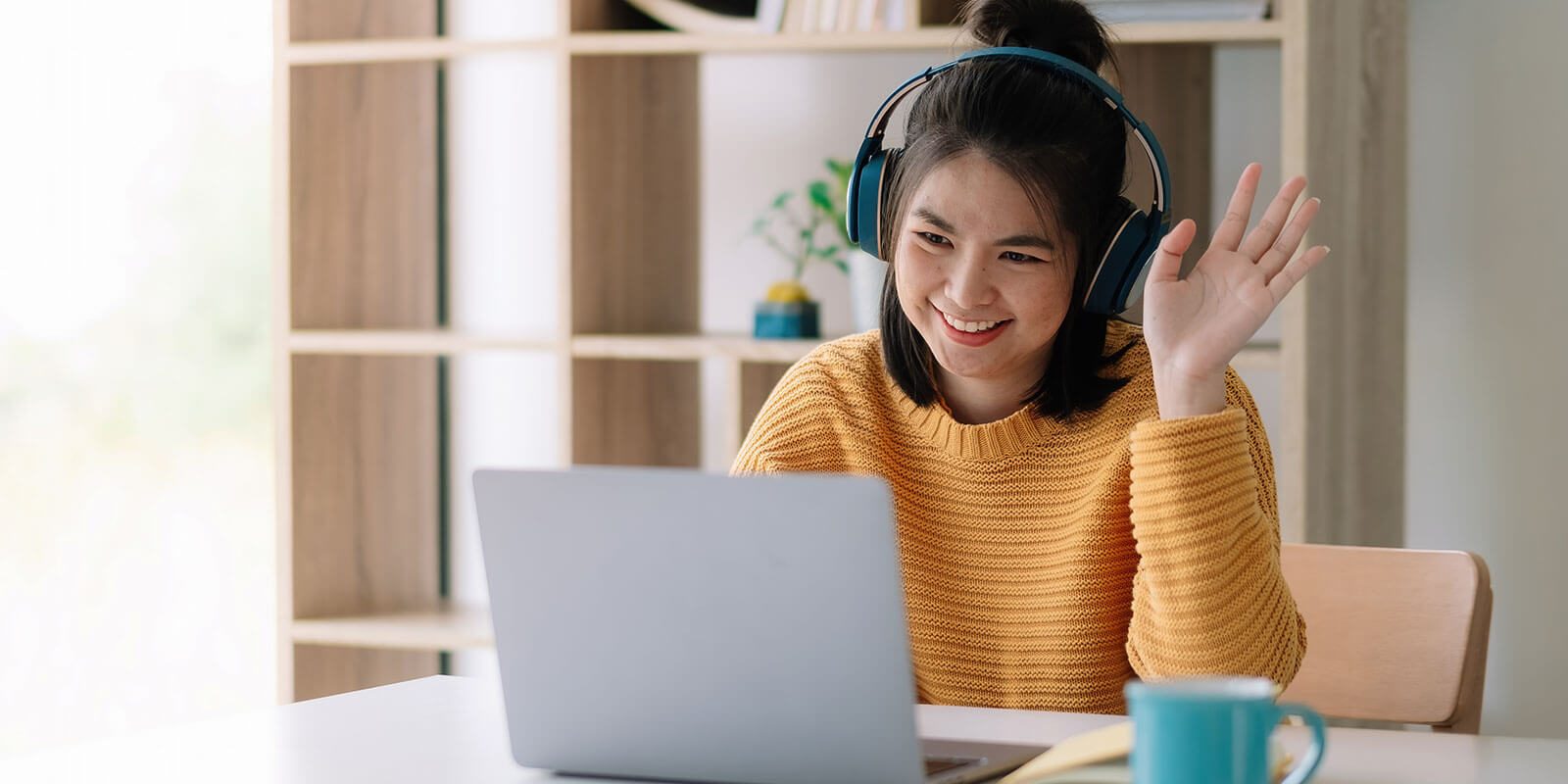 Female student wearing headphones is smiling and waving at her computer