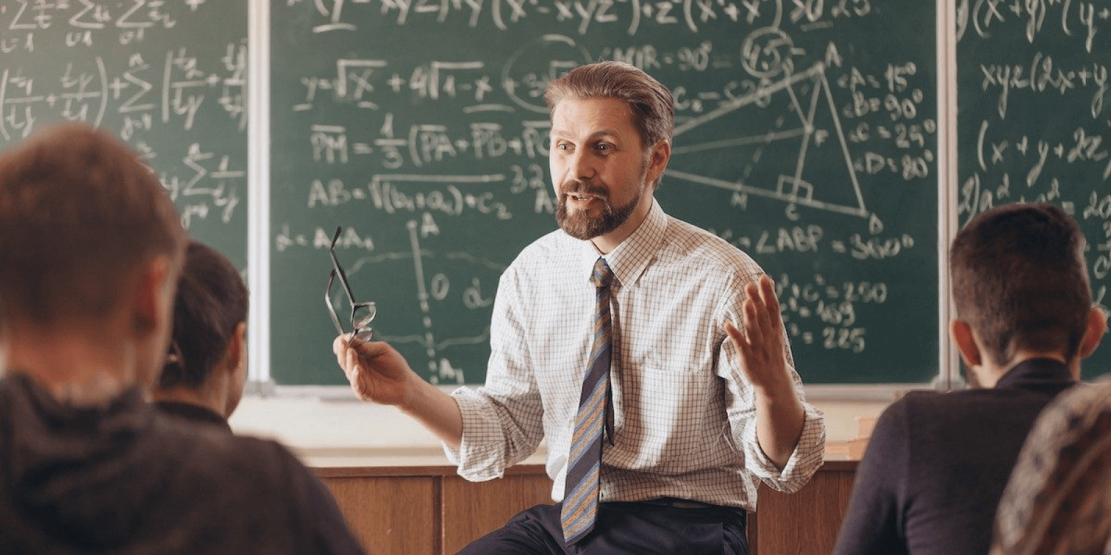 Male teacher is talking to students and is seated at the front of a classroom with mathematical equations and figures on the chalkboard behind him